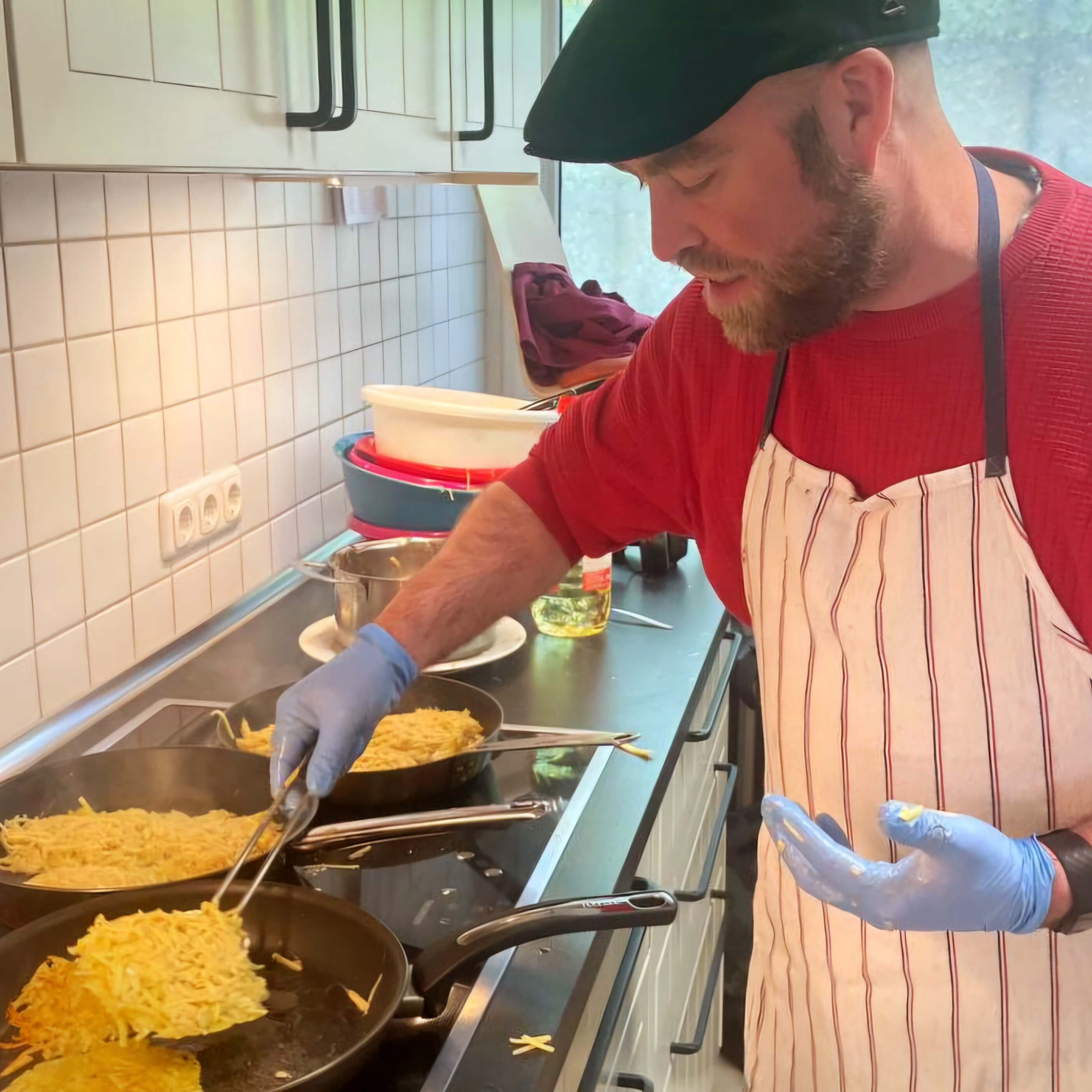 Björn Thoroe trägt eine Schürze und Einmalhandschuhe, während er an einem Herd steht und Kartoffelpuffer brät.