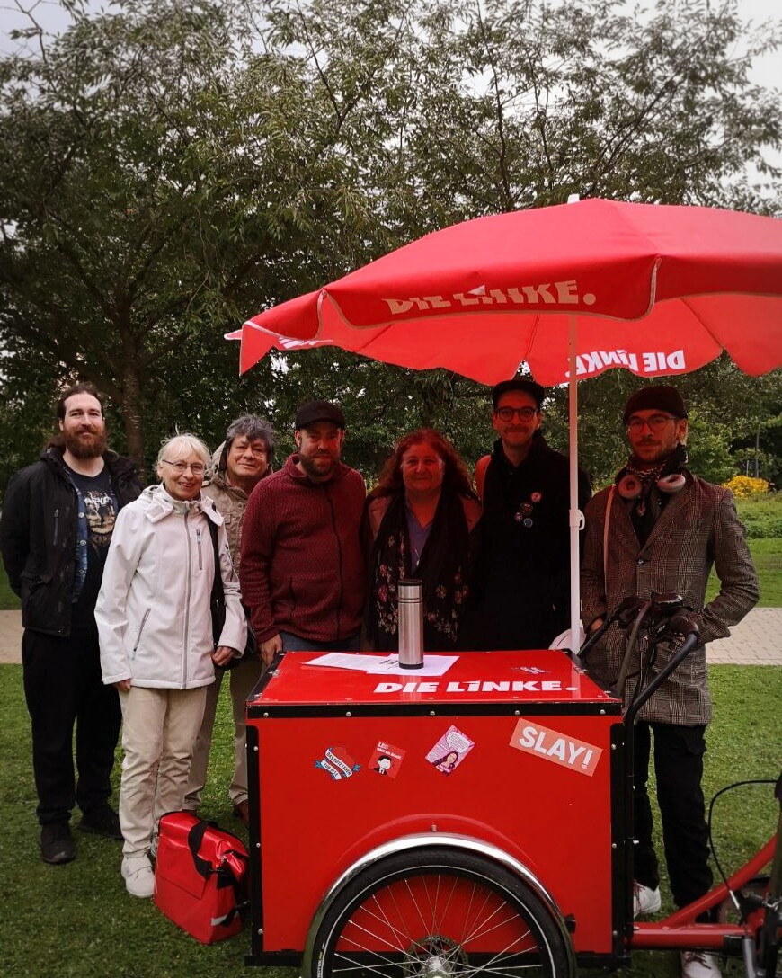 Eine Gruppe von Personen, darunter Ayşe Fehimli und Björn Thoroe, stehen hinter einem roten Lastenfahrrad mit rotem Sonnenschirm, auf welchem "Die Linke" steht.