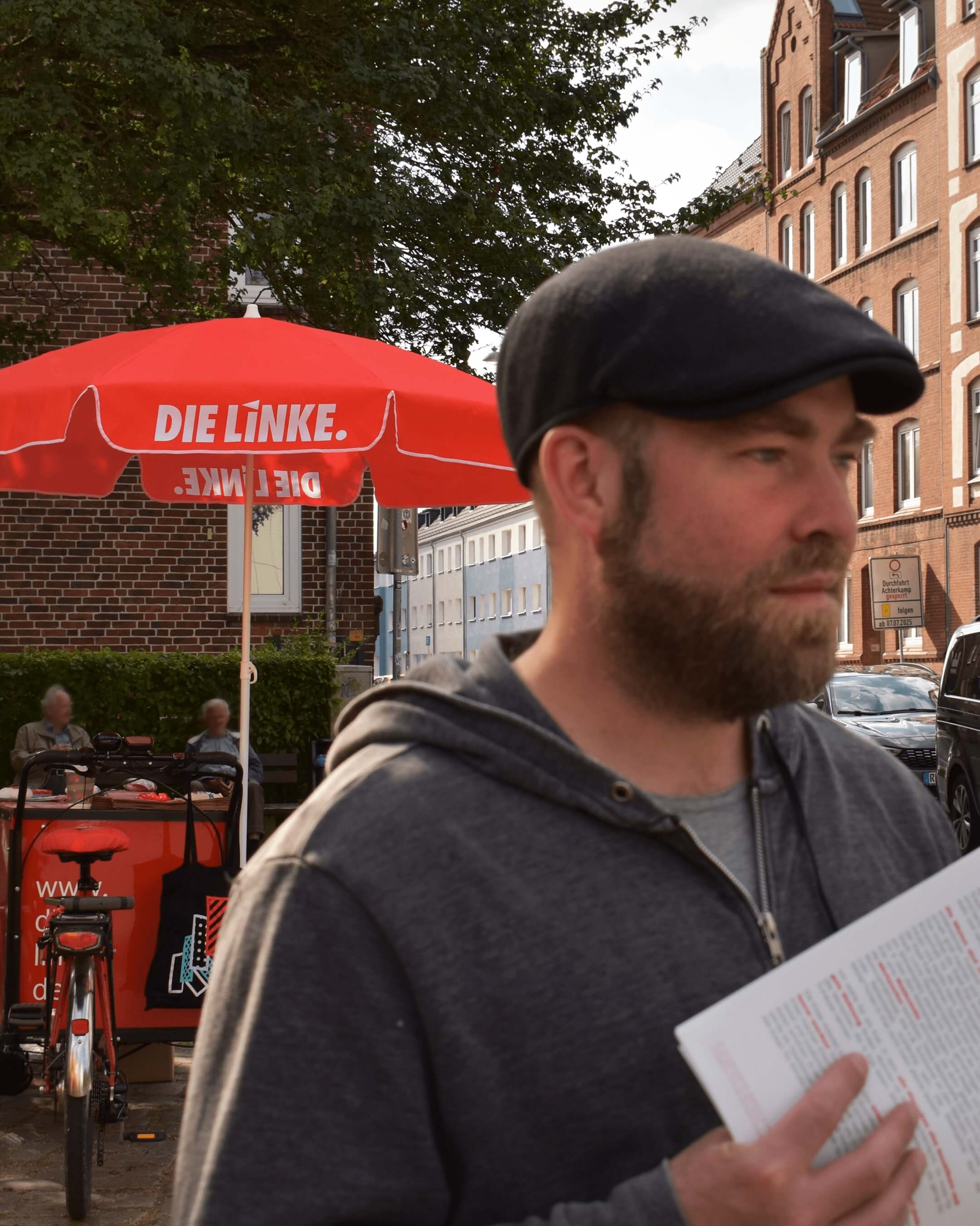 Björn Thoroe im Gespräch. Im Hintergrund ein rotes Lastenfahrrad sowie ein roter Sonnenschirm mit Aufschrift "Die Linke".