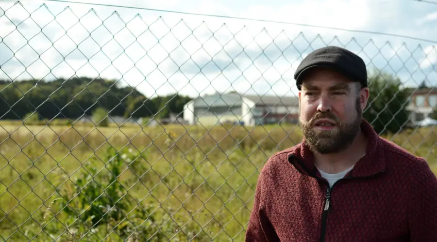 Björn Thoroe mit nachdenklichem Gesichtsausdruck vor einem Maschendrahtzaun, dahinter sind in der Ferne Gebäude auf dem MFG5-Gelände zu sehen.