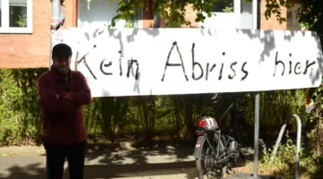 Björn Thoroe steht mit verschränkten Armen vor einem betroffenen Mehrfamilienhaus, vor dem ein Transparent mit folgender Aufschrift hängt: Kein Abriss hier!