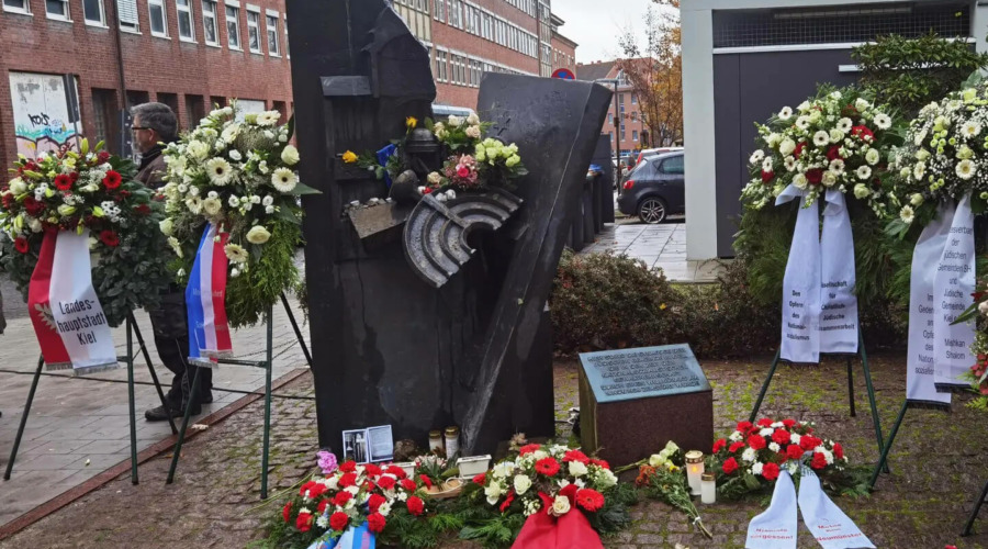 Zu sehen ist der Gedenkstein für die zerstörte Synagoge in der Kieler Goethestraße. Der Gedenkstein ist mit zahlreichen Kränzen umringt, auf dem Gedenkstein liegen Blumen und Steine.