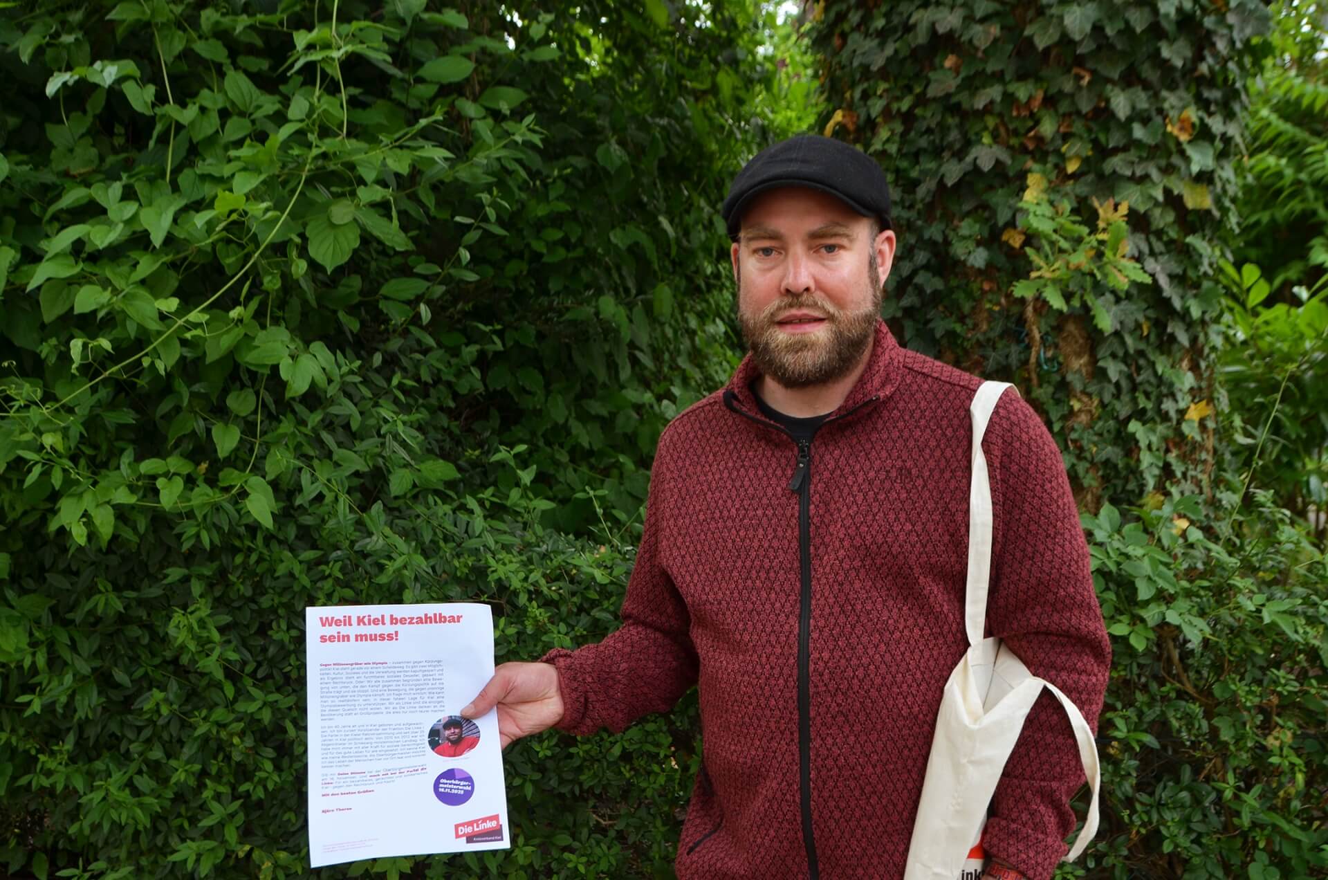 Björn Thoroe hält ein Flugblatt mit dem Titel "Weil Kiel bezahlbar sein muss!" in der Hand, auf welchem er selbst abgebildet ist.