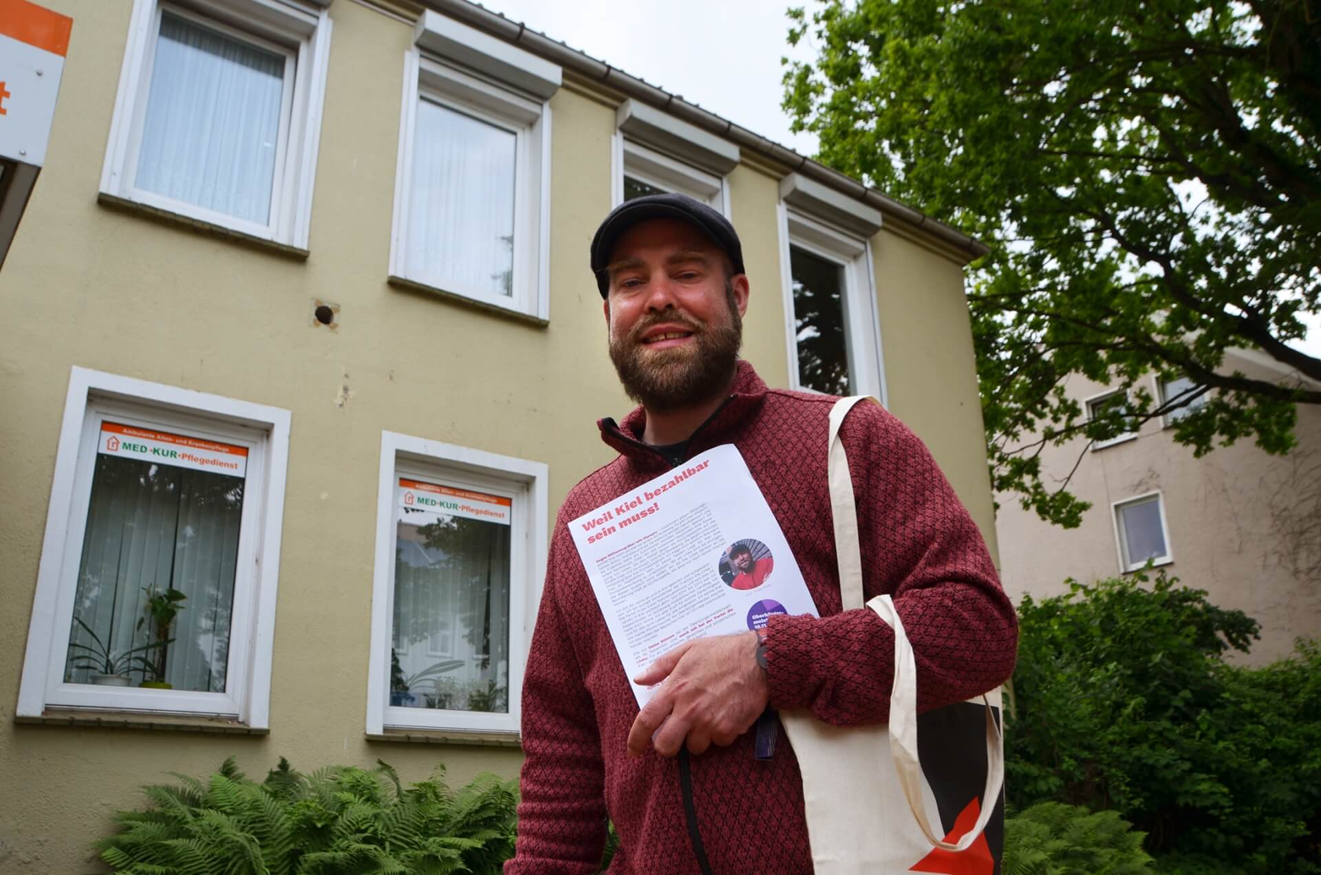 Björn Thoroe lächelt in die Kamera und hält einen Flyer mit dem Title "Weil Kiel bezahlbar sein muss!" in der Hand, auf dem er abgebildet ist.
