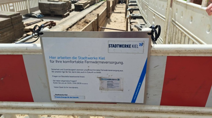An der Absperrung um eine Baugrube hängt ein Schild, auf dem unter anderem folgendes steht: "Hier arbeiten die Stadtwerke Kiel für Ihre komfortable Fernwärmeversorgung."