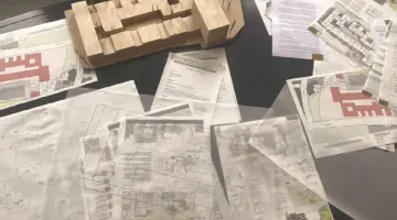 Zu sehen sind zahlreiche Planungs-Skizzen, Risszeichnungen sowie ein hölzernes Gebäude-Modell auf einem Schreibtisch.