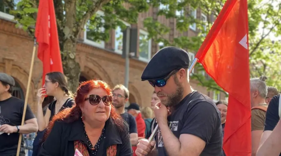 Ayşe Fehimli und Björn Thoroe stehen während einer Demonstration nebeneinander und und unterhalten sich. Björn träge eine rote Fahne, Ayşe hat Flyer in der Hand.