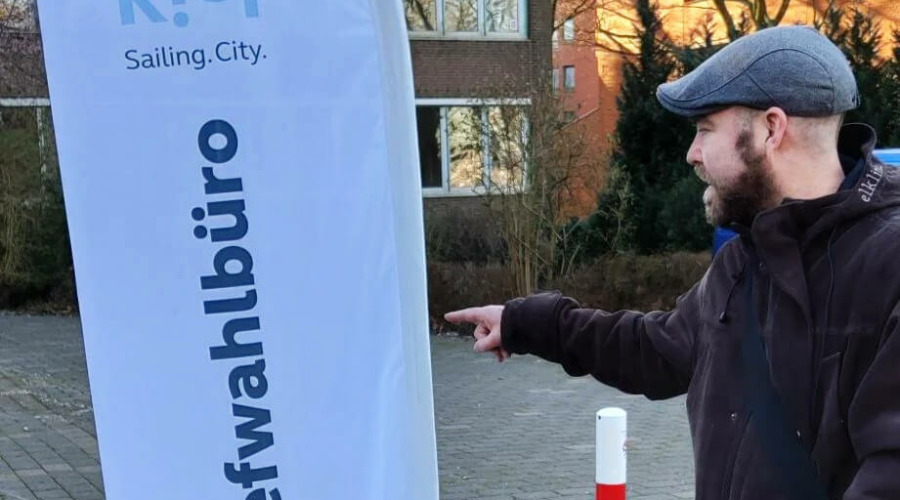 Björn Thoroe zeigt auf ein Schild der Landeshauptstadt Kiel, auf dem "Briefwahlbüro" geschrieben steht.