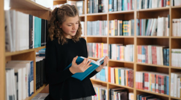 Ein Mädchen steht in einer Bücherei an ein Bücherregal gelehnt und liest in einem Buch.