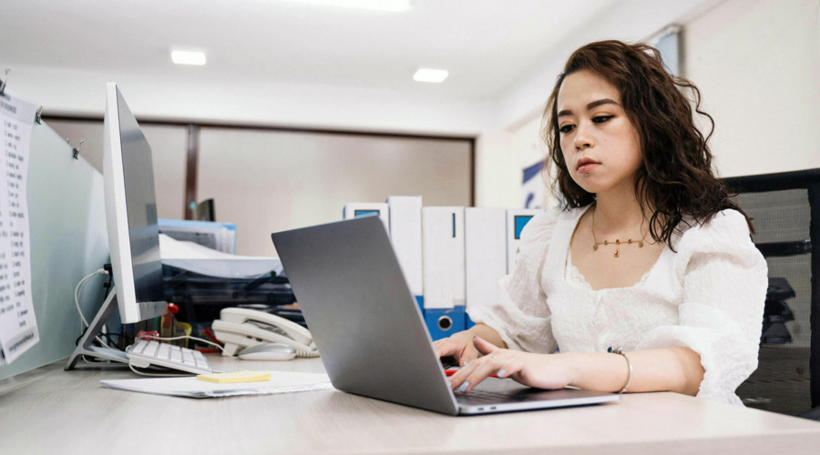 Eine Frau sitzt an einem Schreibtisch-Arbeitsplatz in einem Büro und bedient einen Laptop.