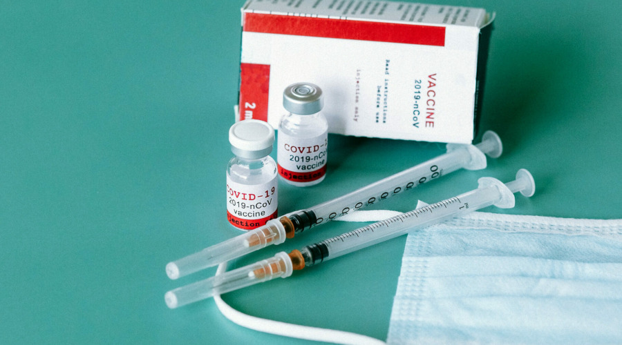 Es sind Fläschchen und eine Packung mit der Aufschrift "Covid-19 2019-nCoV Vaccine" zu sehen, daneben zwei Spritzen und eine Maske.