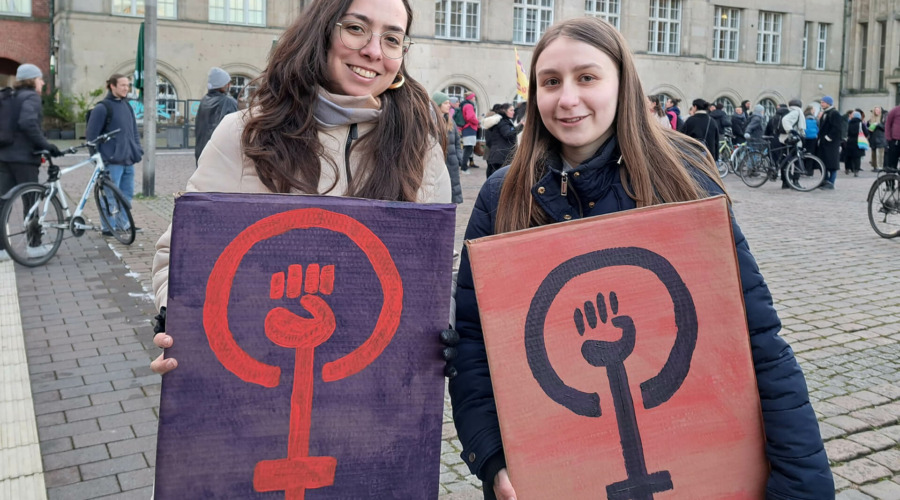 Tamara Mazzi und eine weitere Frau halten auf dem Kieler Rathausplatz Schilder mit dem Feminismus-Symbol.