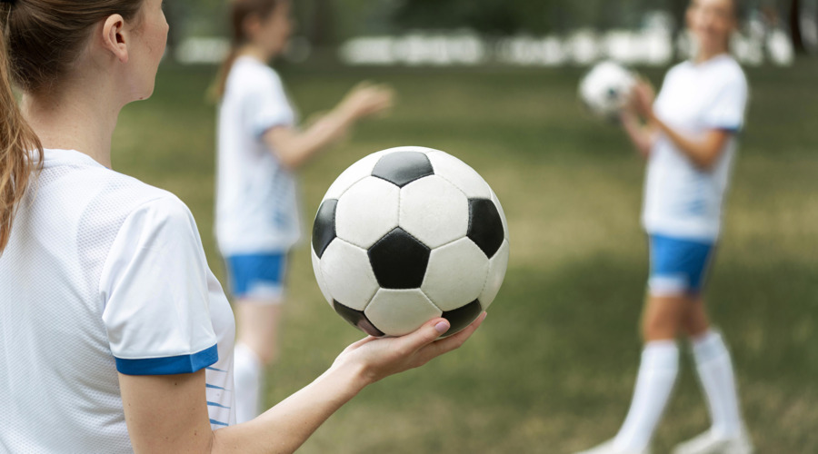 Eine Frau in Trikot hält einen Fußball in der Hand. Im Hintergrund sind weitere Frauen in Trikot auf einem Fußballplatz zu sehen.