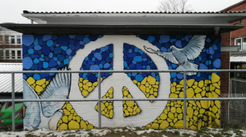 Auf einer Mauer ist das Peace-Zeichen sowie zwei weiße Friedenstauben aufgemalt, eingebettet in die blau-gelben ukrainischen Nationalfarben.