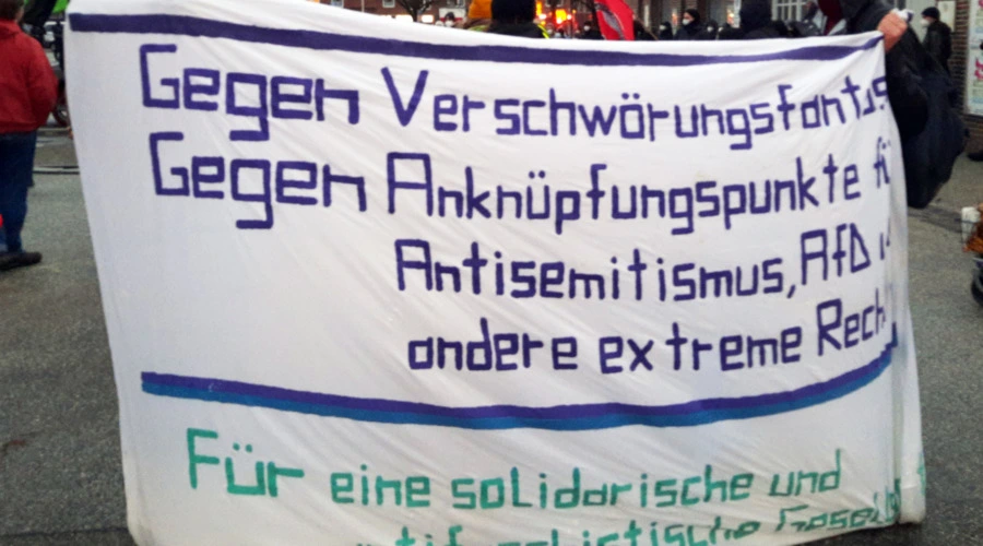 Demonstrierende tragen ein Transparent mit folgender Aufschrift: Gegen Verschwörungsfantasien! Gegen Anknüpfungspunkte für Antisemitismus, AfD und andere extreme Rechte! Für eine solidarische und antifaschistische Gesellschaft!