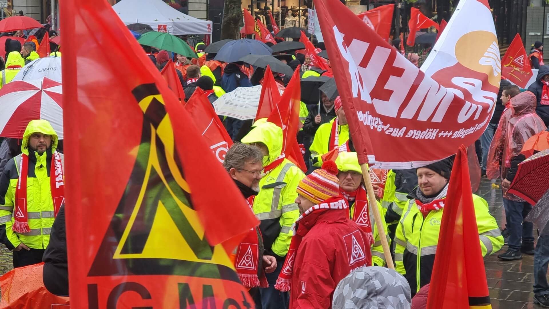 Zu sehen ist eine Demonstration der IG Metall mit zahlreichen roten Fahnen mit dem entsprechenden Gewerkschaftslogo bei Regenwetter.