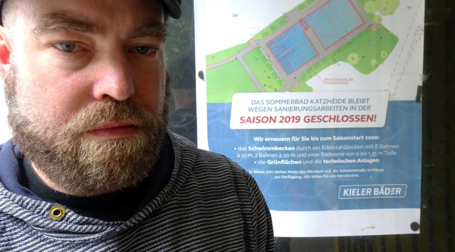 Björn Thoroe vor dem geschlossenen Freibad Katzheide. Im Hintergrund ein Schild mit folgender Aufschrift: "Das Sommerbad Katzheide bleibt wegen Sanierungsarbeiten in der Saison 2019 geschlossen!"