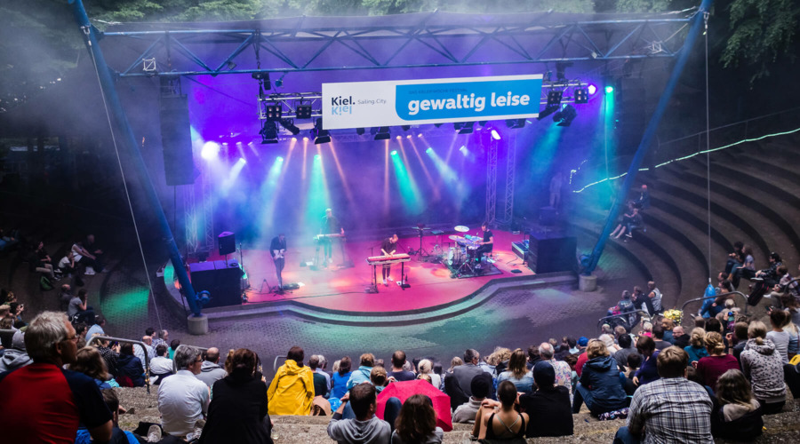 Ein Konzert in der Freilichtbühne Kiel. Auf den Rängen sitzen zahlreiche Zuschauer, auf der Bühne spielt eine Band. Über den Bühne ist ein Schild mit der Aufschrift "Kiel. Sailing City. Gewaltig leise" befestigt.