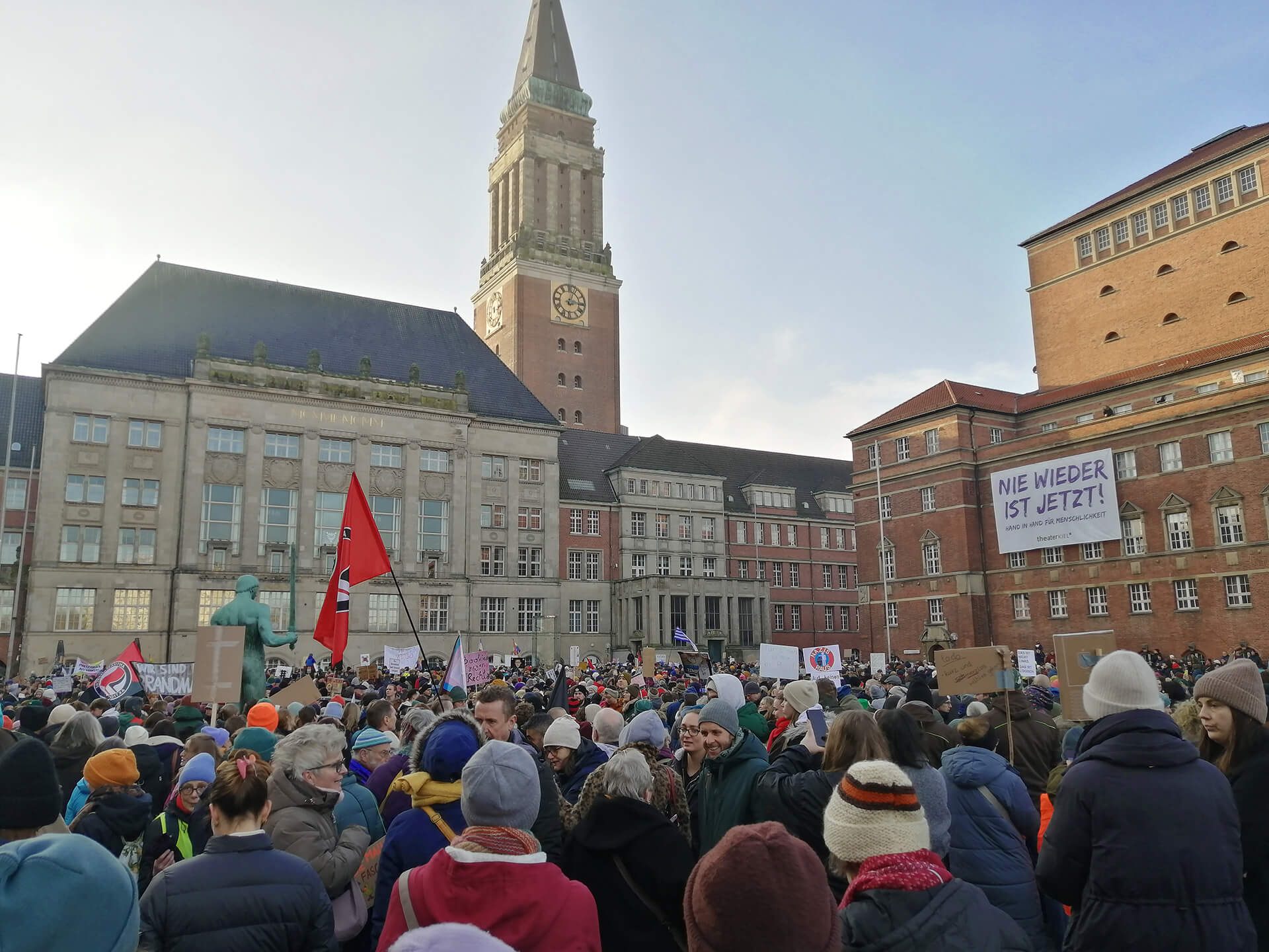 Eine demonstrierende Menschenmenge auf dem Rathausplatz in Kiel. In der Mitte des Fotos ist der markante Rathausturm zu sehen. Am Theatergebäude rechts im Bild hängt ein großes Transparent mit der Aufschrift "Nie wieder ist jetzt!"