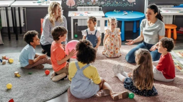 Eine Gruppe kleiner Kinder sitzt mit zwei Erzieherinnen in einem Kreis. Sie spielen mit einem Ball. Im Umfeld ist die Einrichtung einer Kindertagesstätte zu sehen.