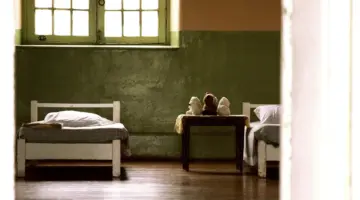 Ein karger Raum mit kaum zu erreichendem Fenster. Es sind zwei Betten zu sehen, in der Mitte ein Nachttisch, auf welche drei Teddybären sitzen.