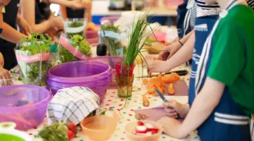 Kinder stehen um einen Tisch herum und bereiten Essen zu. Auf dem Tisch stehen verschiedene Kräuter, ein Kind hat ein kleines Küchenmesser in der Hand, ein anderes schält eine Möhre.