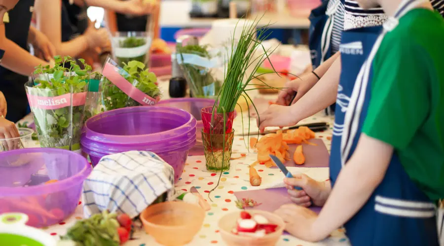 Kinder stehen um einen Tisch herum und bereiten Essen zu. Auf dem Tisch stehen verschiedene Kräuter, ein Kind hat ein kleines Küchenmesser in der Hand, ein anderes schält eine Möhre.