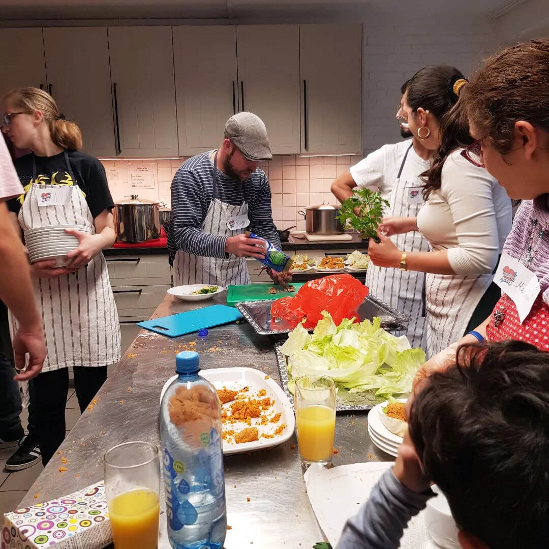 Mehrere Personen in einer Küche, bei der Vorbereitung von Mahlzeiten. Darunter Tamara Mazzi und Björn Thoroe.