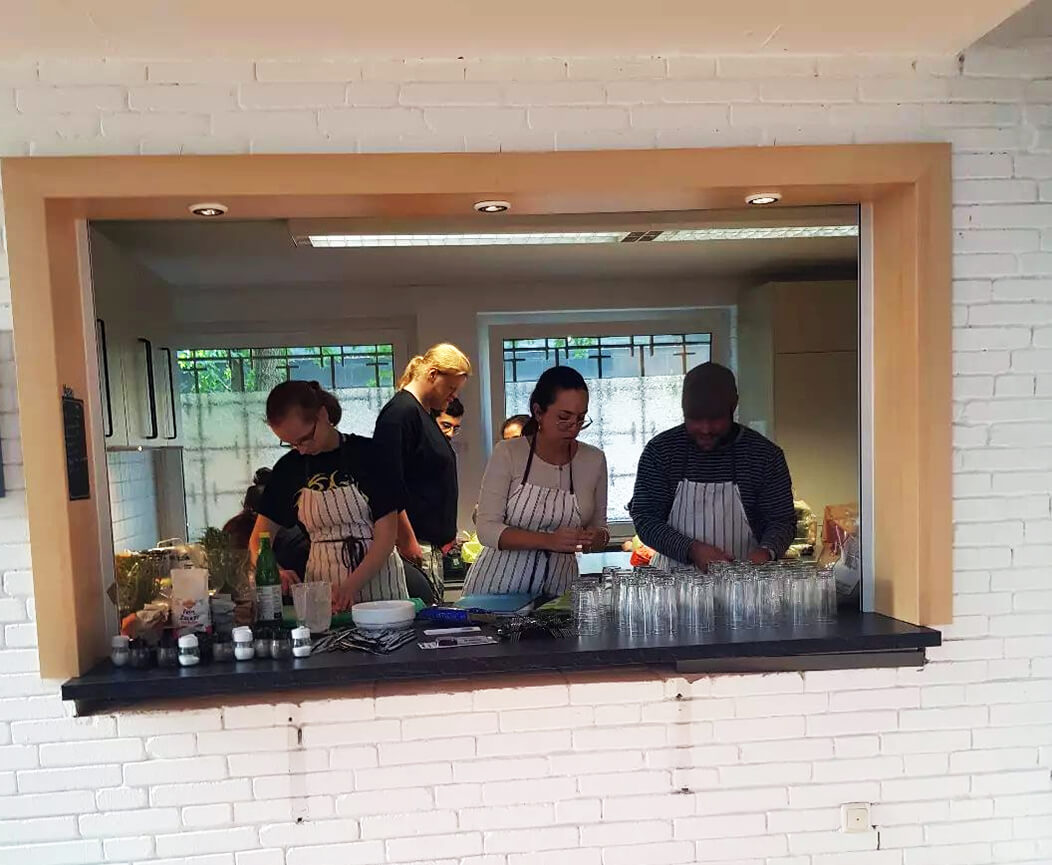 Eine Gruppe von Personen bereitet in einer Küche Mahlzeiten zu, darunter Tamara Mazzi und Björn Thoroe. Zu sehen ist diese Szene durch eine Durchreiche, auf der u.a. viele noch leere Gläser stehen.