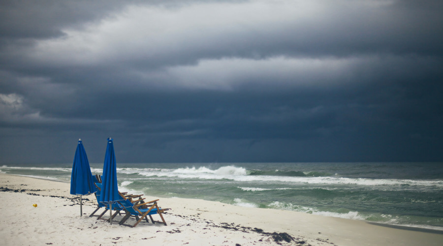 Blick vom Strand auf ein aufgewühltes Meer mit Brandungs-Wellen. Über dem Meer tief hängende, dunkle Wolken. Auf dem Strand stehen einige Liegestühle und zwei Sonnenschirme in blauer Farbe.