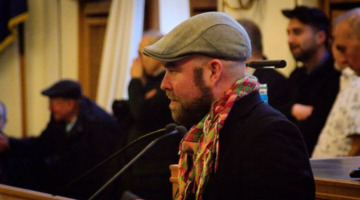 Björn Thoroe steht am Redepult im Kieler Ratssaal und trägt dabei ein Halstuch in kurdischen Farben.