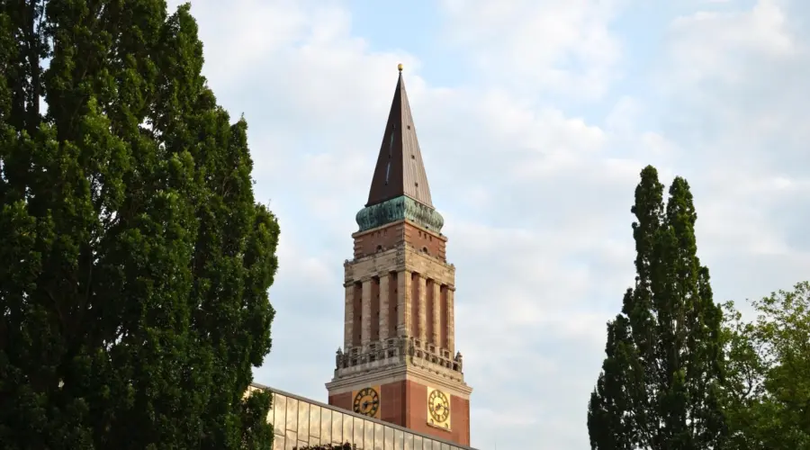 Zu sehen ist der markante Rathausturm im Kiel, davor die gläserne Fassade des Theatergebäudes. Im Vordergrund Bäume mit grünen Blättern.