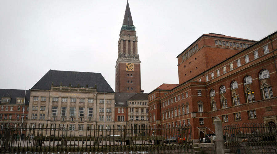 Zu sehen sind das Kieler Rathaus mit dem markanten Rathausturm und das Kieler Opernhaus. Auf einer Mauer vor dem Rathausplatz ist der Schriftzug "Revolutionsstadt" aufgesprüht.