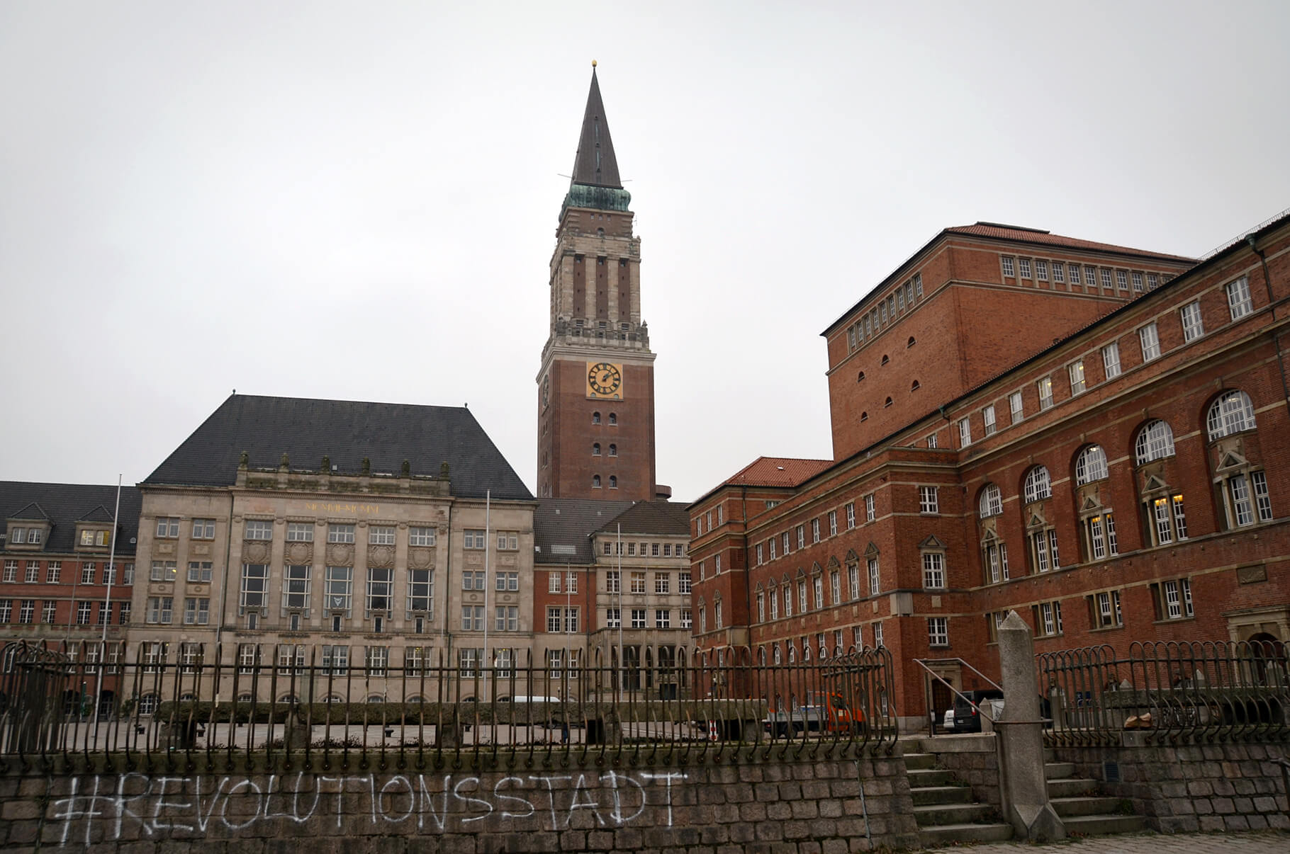 Zu sehen sind das Kieler Rathaus mit dem markanten Rathausturm und das Kieler Opernhaus. Auf einer Mauer vor dem Rathausplatz ist der Schriftzug "Revolutionsstadt" aufgesprüht.