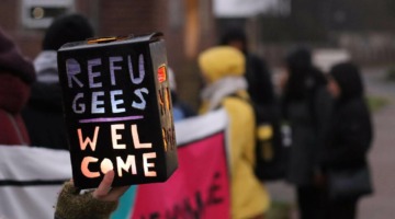 Vor einer Demonstration mit Transparent wird mit einer behandschuhten Hand eine kleine Laterne gehalten, welche die Aufschrift "Refugees Welcome" trägt.
