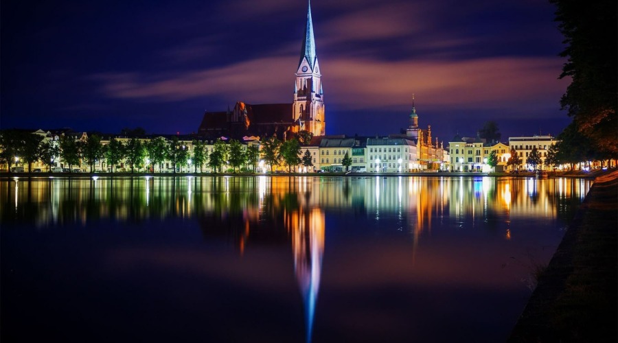Zu sehen ist die hell erleuchtete Innenstadt von Schwerin bei Nacht. Im Zentrum des Fotos ist der angestrahlte Schweriner Dom, welcher sich auch auf einer Wasserfläche am unteren Bildrand spiegelt.