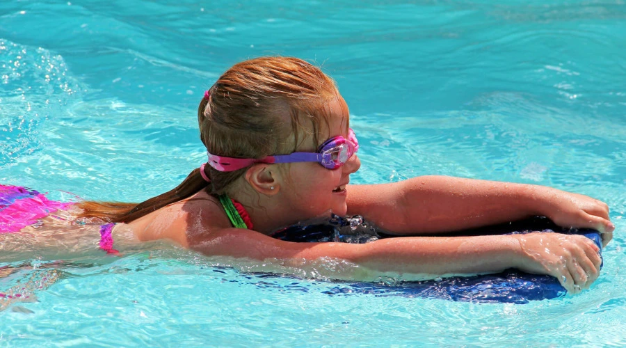 Ein junges Mädchen mit Schwimmbrille schwimmt gestützt auf ein Schwimmbrett durchs Wasser.