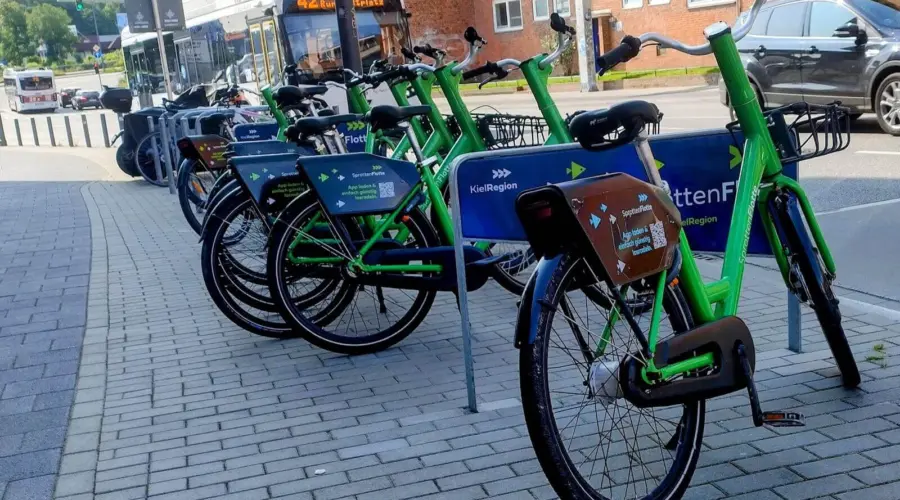 Eine Station des Bike-Sharing-Systems "Sprottenflotte" mit zahlreichen Leihrädern. Im Hintergrund die Alte Lübecker Chaussee in Kiel, mit Straßenverkehr und einem vorbeifahrendem Bus der Linie 42.