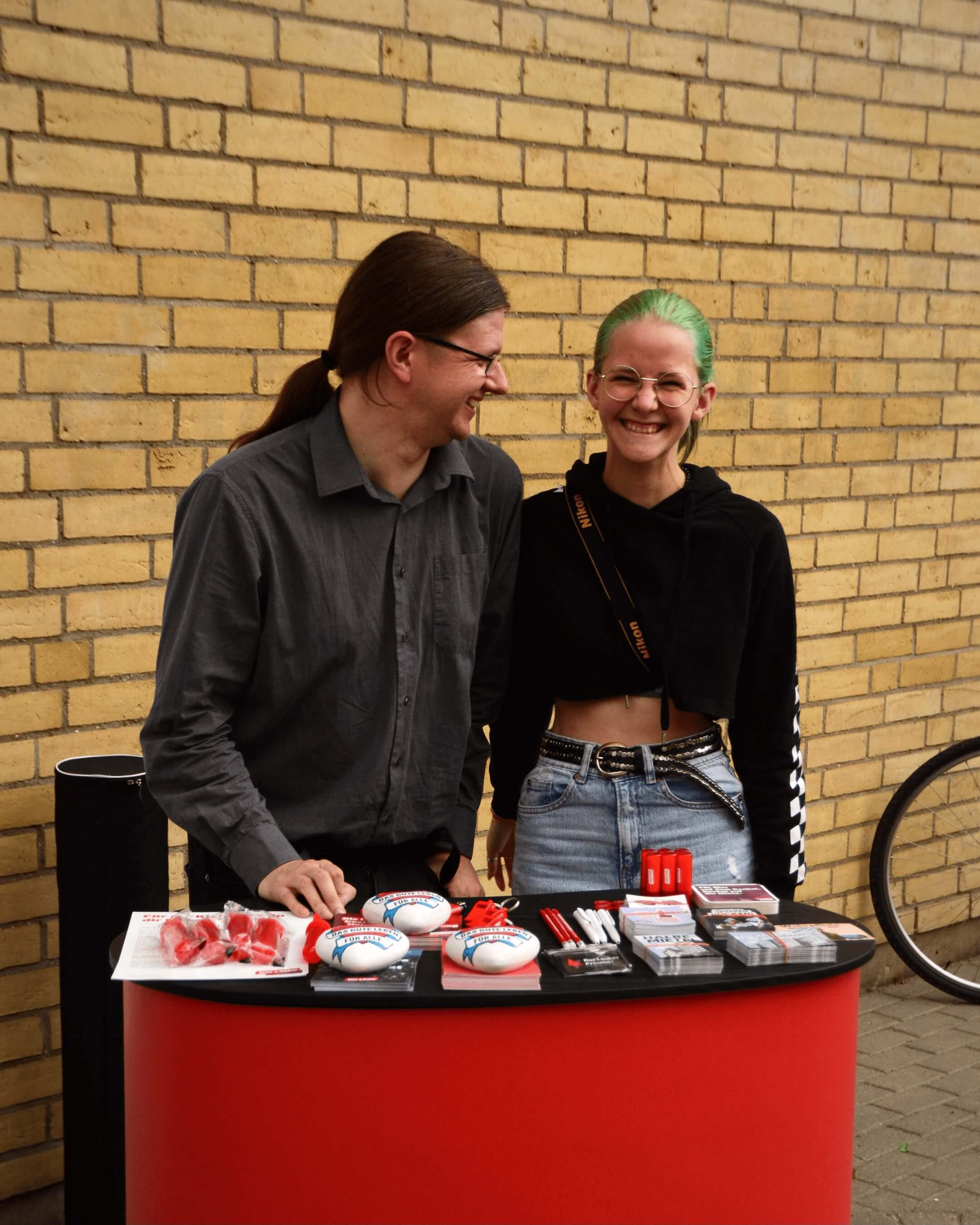 Ein Mann und eine Frau mit grünen Haaren stehen hinter einem Infostand von "Die Linke".