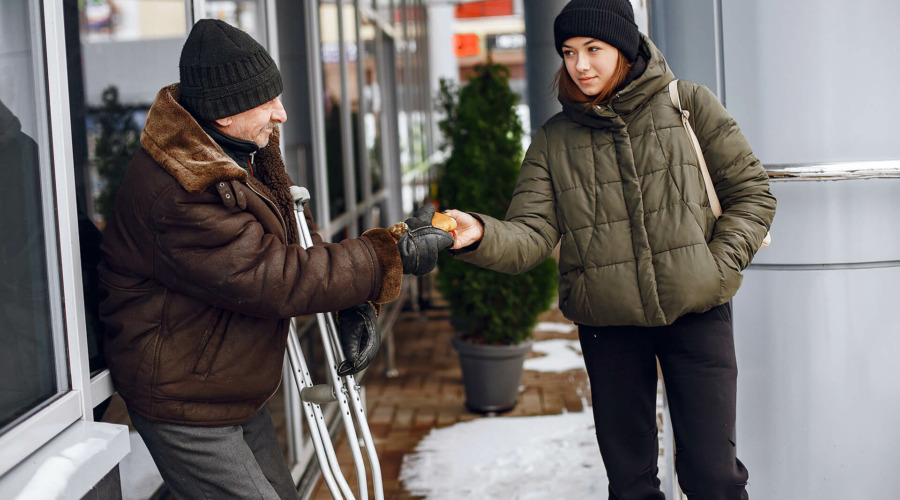 Eine junge Streetworkerin reicht einem älteren Mann mit Krücken etwas. Die Szene spielt sich auf einem verschneiten Gehweg im Winter ab.