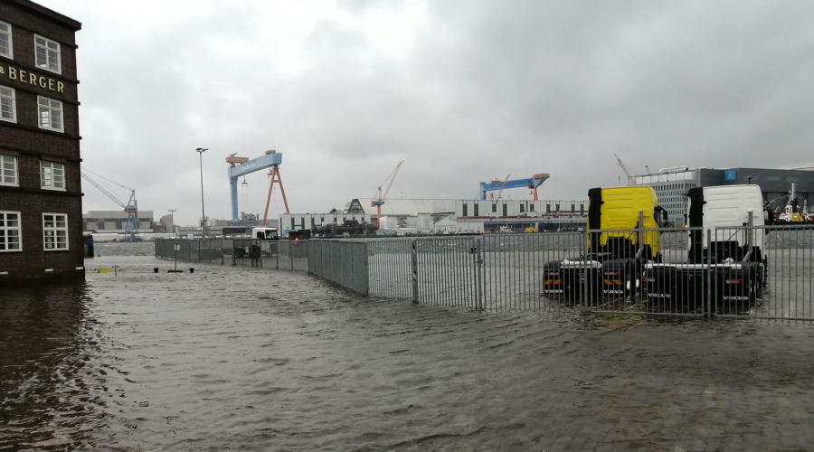 Zu sehen ist der überflutete Sartori-Kai im Kieler Hafen während der Sturmflut am 20. Oktober 2023. Im Hintergrund die bekannten blauen Kräne der Werft. Links im Bild zwei bereits im Wasser stehende LKW-Zugmaschinen.