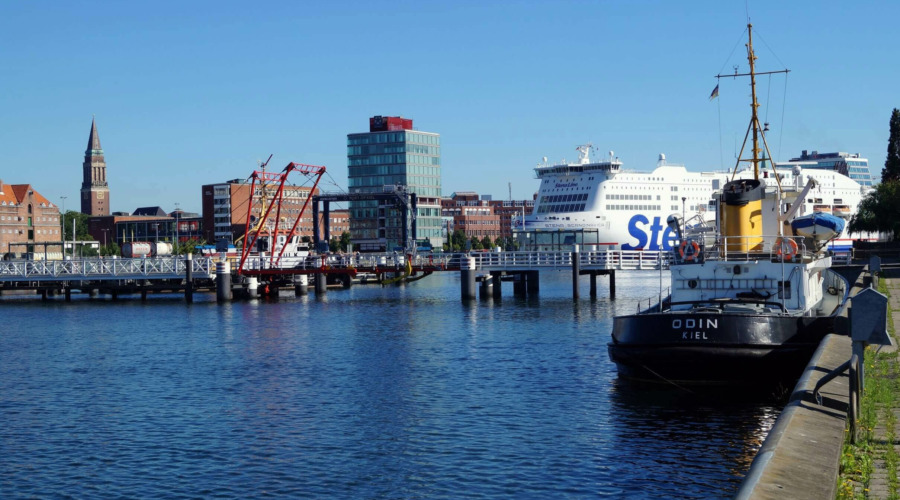 Der Kieler Hafen, im Hintergrund sind eine Fähre sowie die Hörnbrücke zu sehen, im Vordergrund ein Schlepper-Schiff.