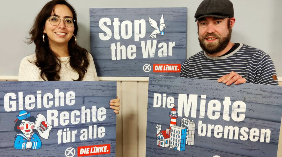 Tamara Mazzi und Björn Thoroe halten Schilder mit folgenden Texten: "Gleiche Rechte für alle. Die Linke" sowie "Die Miete bremsen. Die Linke". Im Hintergrund steht ein Schild mit der Aufschrift "Stop the War. Die Linke."
