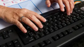 Zwei Hände tippen auf einer Tastatur. Unter der Tastatur ist ein Kalender zu sehen.