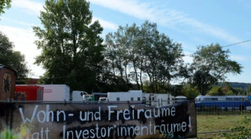 Im Hintergrund sind verschiedene Wagen, ein alter Bus und LKW zu sehen. Im Vordergrund hängt an einem Bauzaun ein Transparent mit folgender Aufschrift: "Wohn- und Freiräume statt Investor*innenträume!"